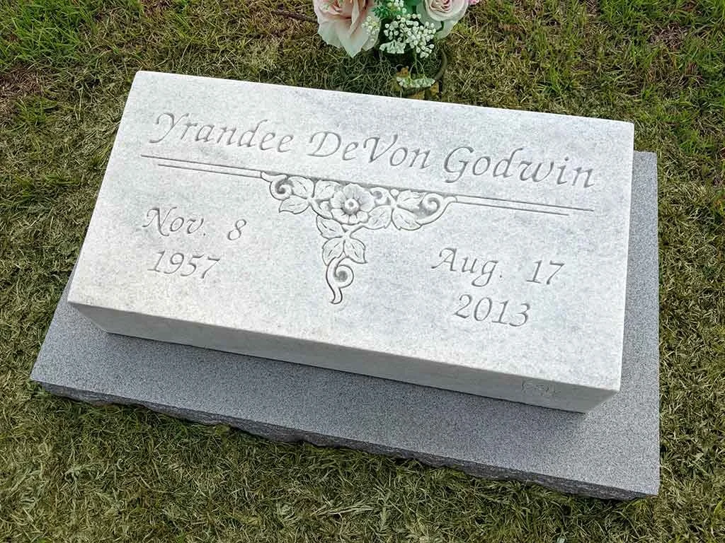 Grave marker with engraved floral decoration, inscribed with the name Trande DeVon Godwin, birth date November 8, 1957, and death date August 17, 2013, placed on a grassy ground with flowers nearby.