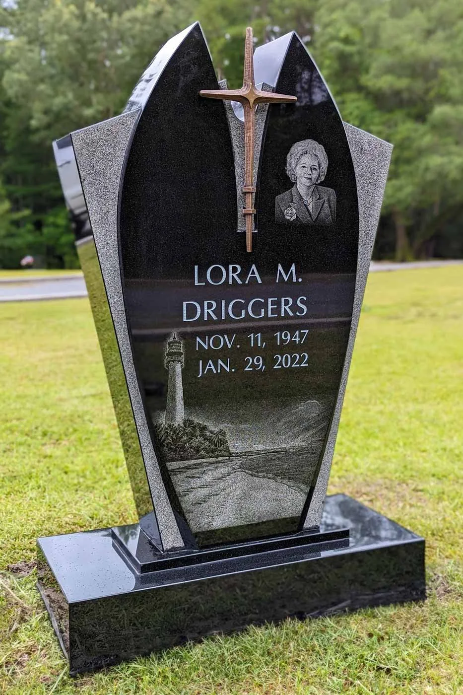 Black granite gravestone with engraved portrait of a woman, a cross with a copper finish on top, and etched lighthouse scene at the bottom, inscribed with the name Lora M. Driggers and birth and death dates.
