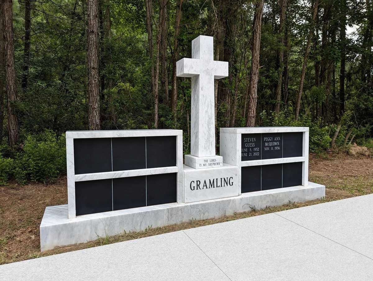 White marble grave marker with a cross, engraved with the name Gramling, and two sections with black plaques. One side has the names and birth and death dates of Steven Guess and Peggy Ann McQuown. The other section has a blank plaque. The background