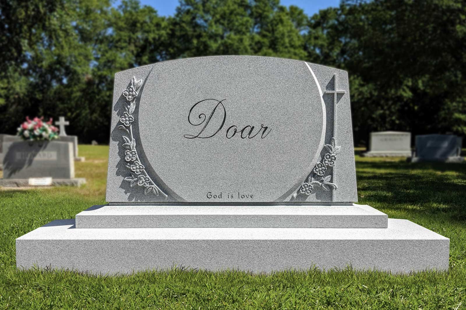 A gray granite headstone in a cemetery with the word 'Doar' inscribed, decorated with floral carvings and a cross, with other gravestones and trees in the background.