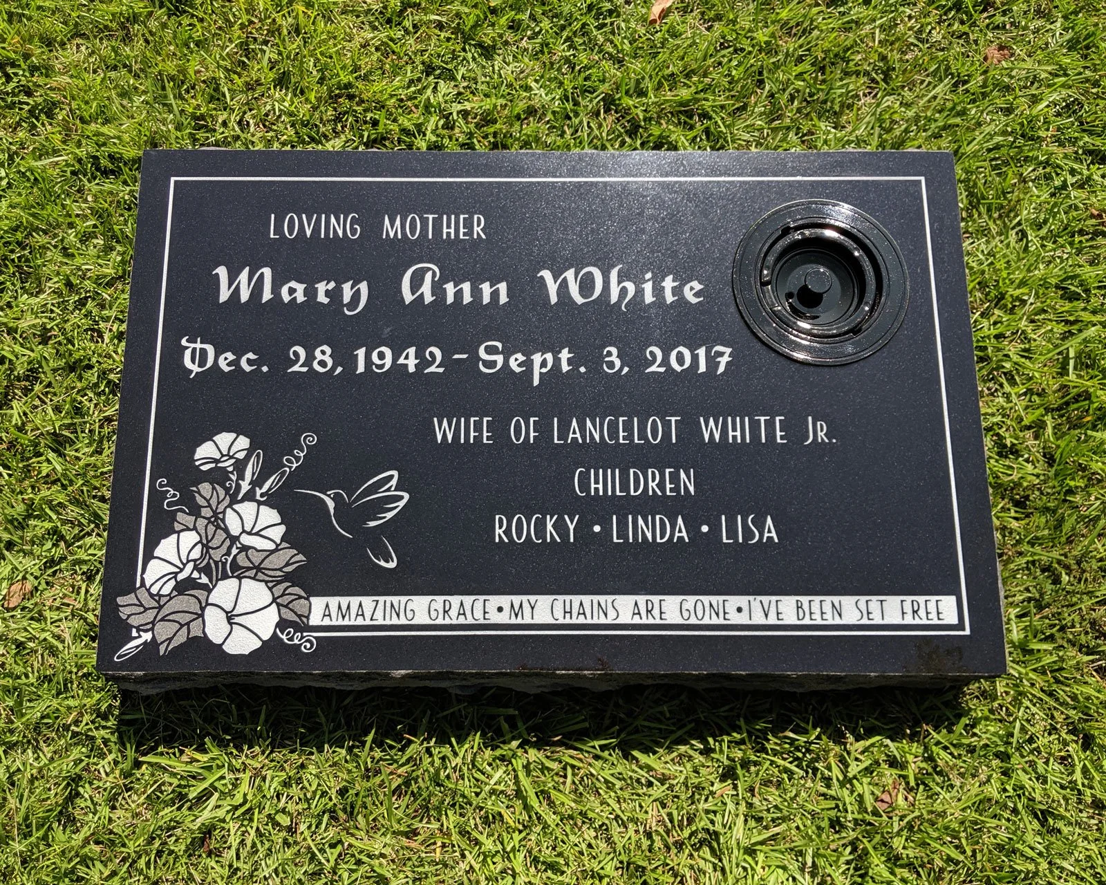 Memorial plaque for Mary Ann White, with flower and hummingbird illustration, set on grass.