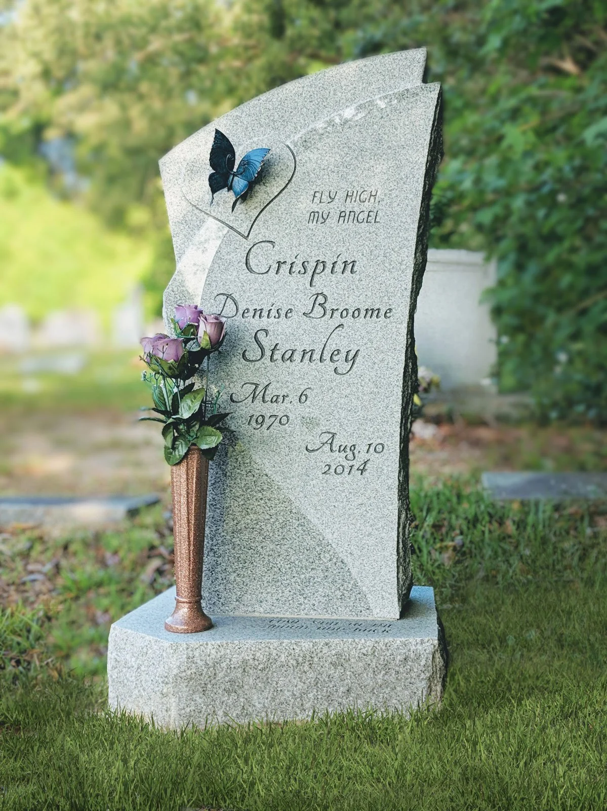 Gray granite headstone with a black and blue butterfly attached at the top, a small flower arrangement in a brown vase, and inscribed with the name Crispin Stanley, born March 6, 1970, and died August 10, 2014, in a grassy area with blurred greenery 
