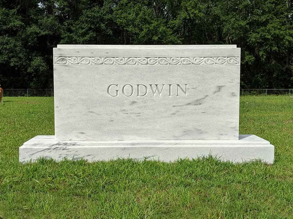 White granite tombstone with the name 'GODWIN' engraved on it, decorated with a carved floral pattern at the top, situated on a grassy area with trees in the background.