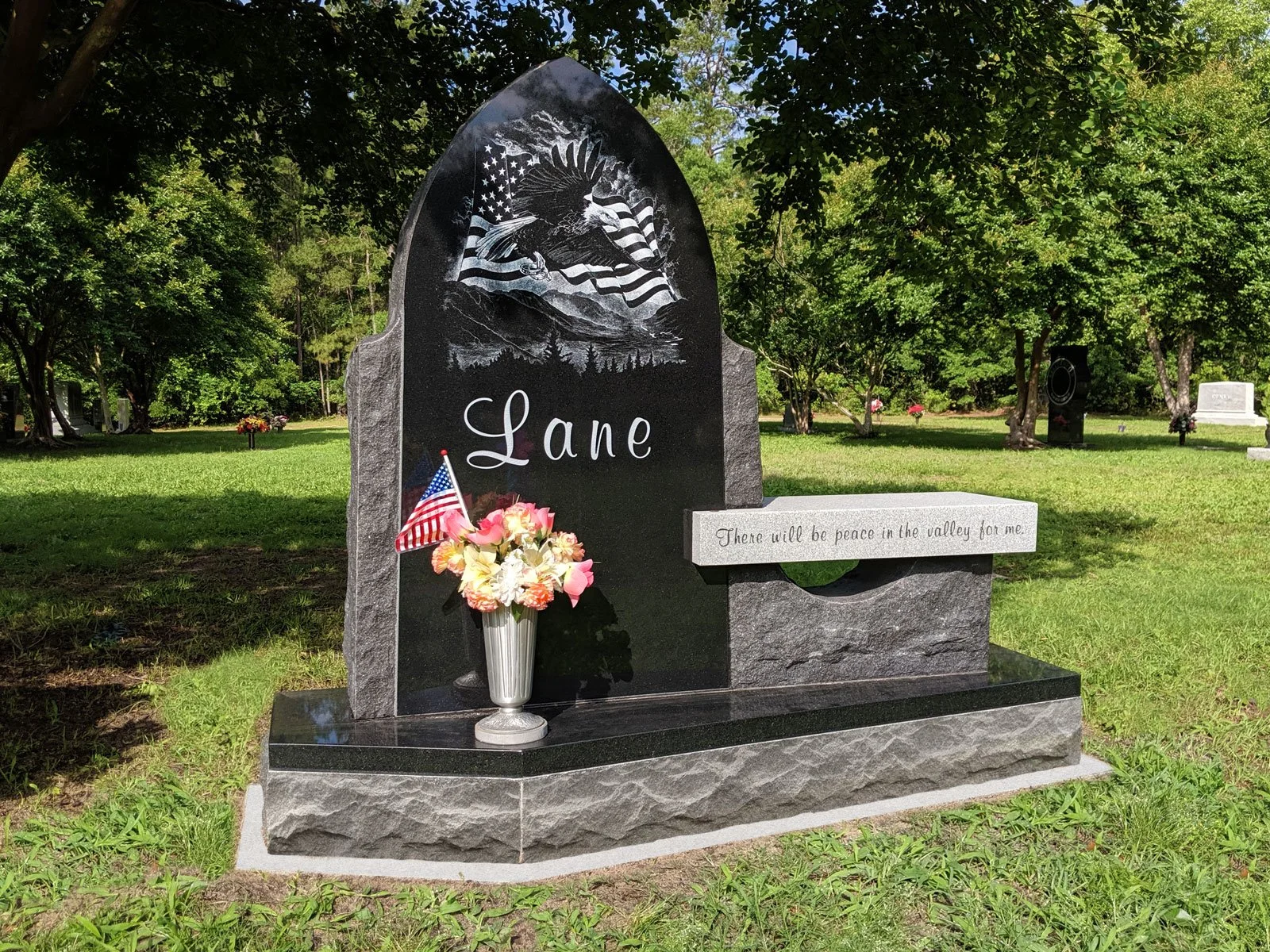 Gravestone with the name 'Lane' engraved, decorated with a bouquet of flowers and American flags, featuring an eagle with an American flag and mountain scenery engraved at the top, in a grassy cemetery with trees.