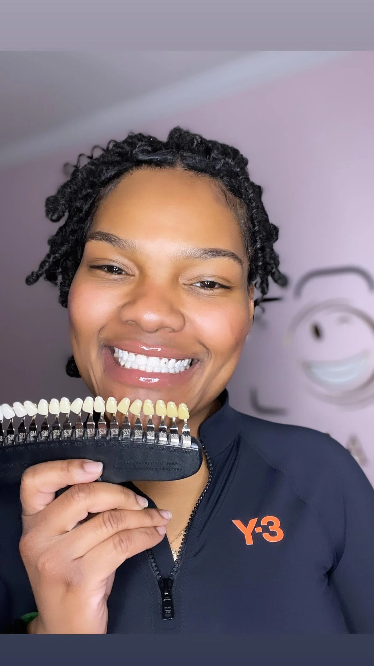 A woman smiling and holding a shade guide for teeth coloring in front of her. She is wearing a black jacket with an orange 'Y-3' logo.