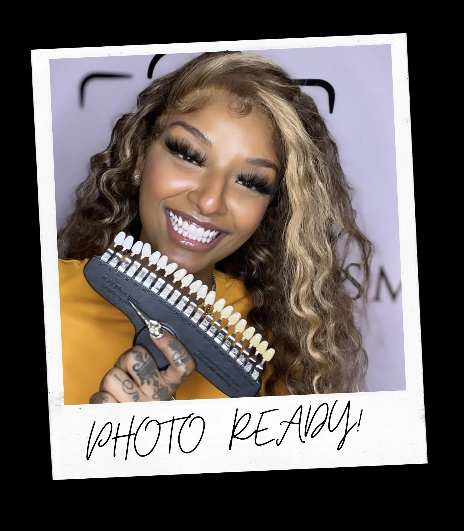 A woman with curly hair holding a gun-shaped makeup palette, smiling broadly at the camera, with a gray background and black text overlay that reads 'PHOTO READY!'