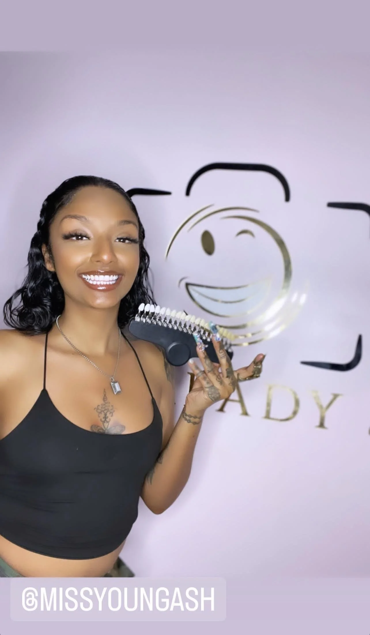 Smiling young woman with tattoos, wearing a black crop top, holding a nail polish fan, standing in front of a wall with a gold and black logo and the text '@MISSYOUNGASH.'