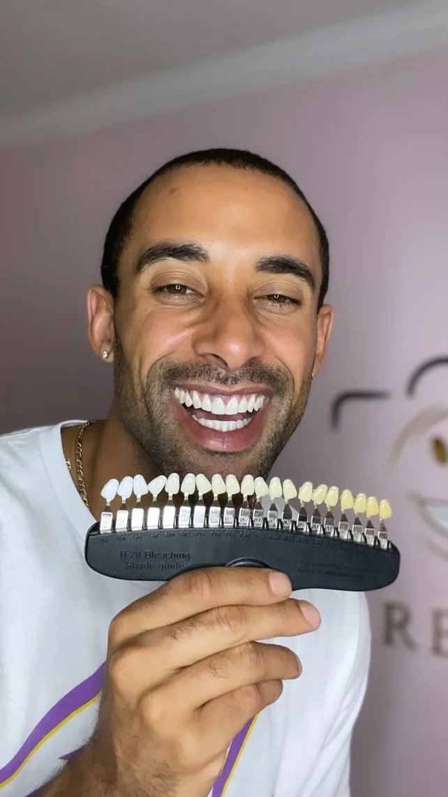 A man smiling while holding a shade guide with tooth color samples. He has short dark hair, a beard, and is wearing a white t-shirt.