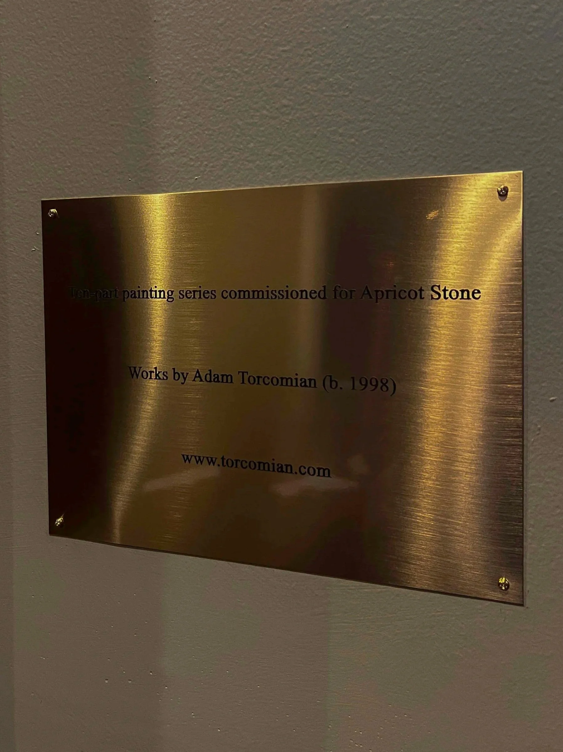 Brass plaque mounted on a wall with engraved text about a painting series and the artist Adam Torcomian.