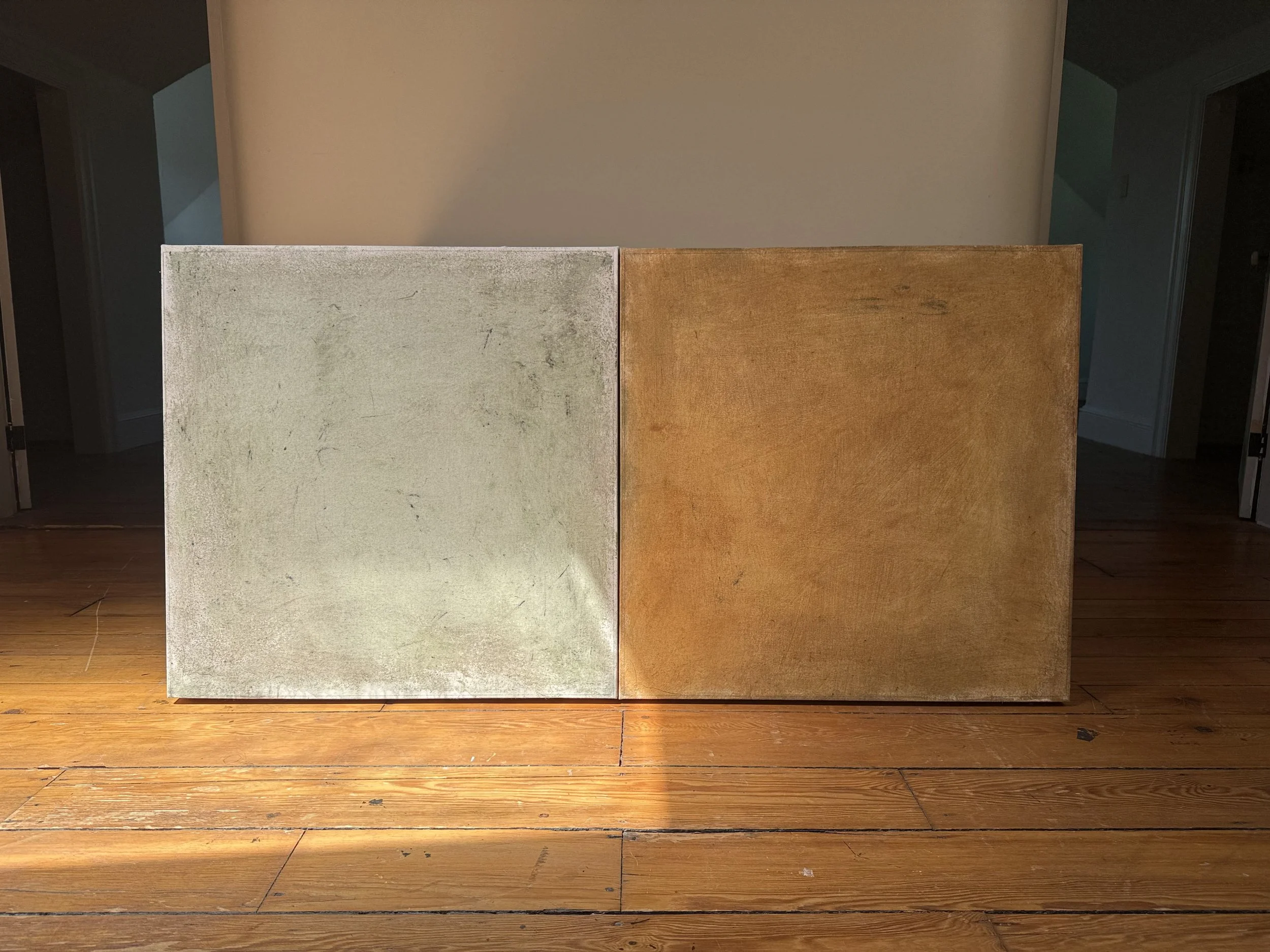 Two panels, one white and weathered, the other brown and wooden, are placed side by side on a wooden floor with a plain wall in the background.