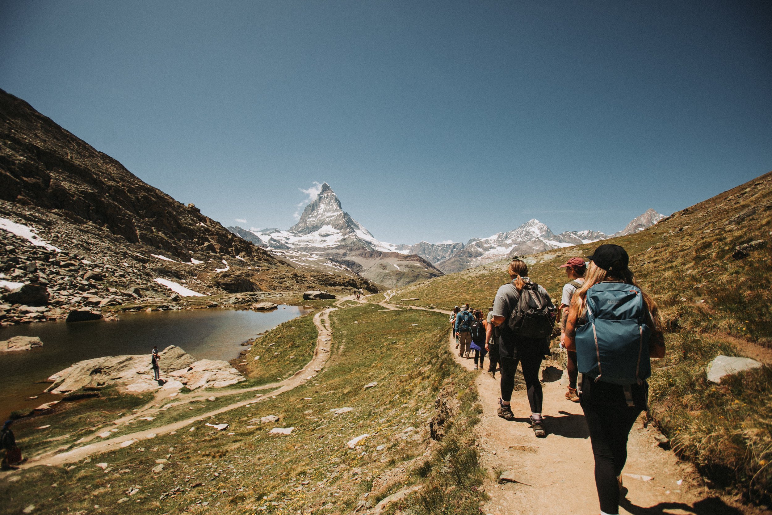 Group of hikers walking on a trail in a mountainous landscape with the Matterhorn mountain in the background, near a small lake and snow-capped peaks.