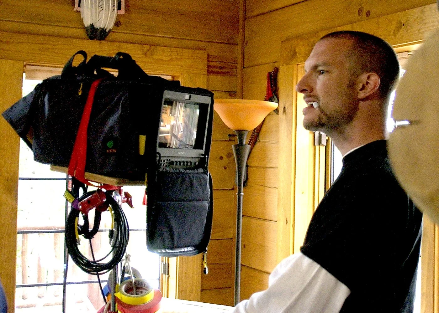 A man with a short beard and short hair is looking at a video camera mounted on a stand inside a wooden cabin. The cabin has wood-paneled walls and a window in the background.