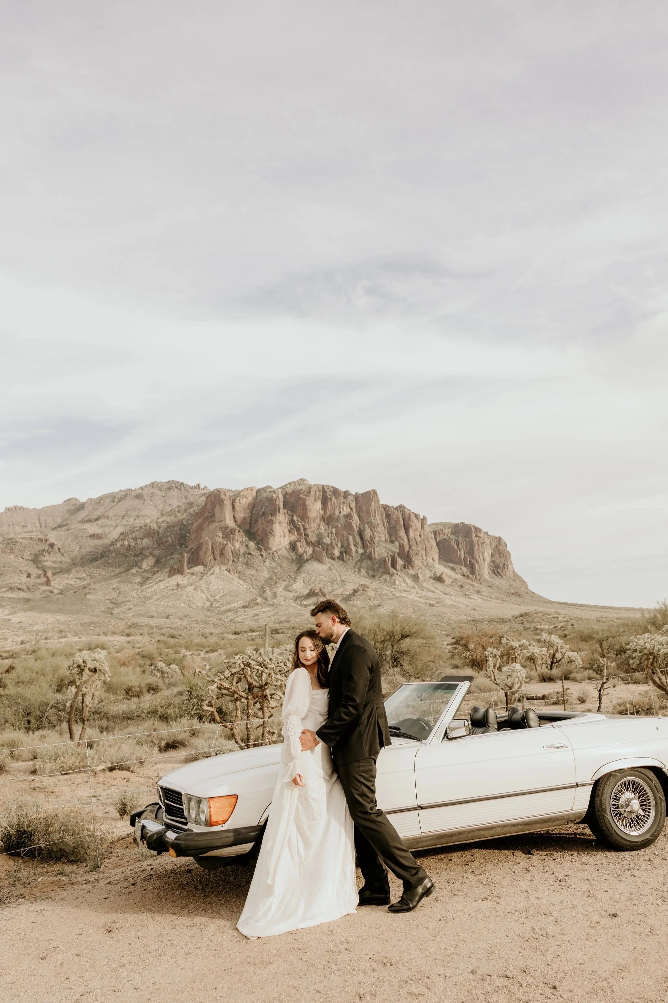 A black and white photograph of a bride and groom sitting on a bench, sharing a kiss. The bride is holding a bouquet of flowers, and holding the groom's hand, with the groom wearing a suit and glasses. The background features a plain wall with decorative paneling and a hanging lantern.