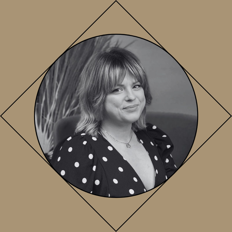 Black and white portrait of a female hairstylist with shoulder-length hair wearing a polka dot dress and a necklace, sitting in front of a background with plants.