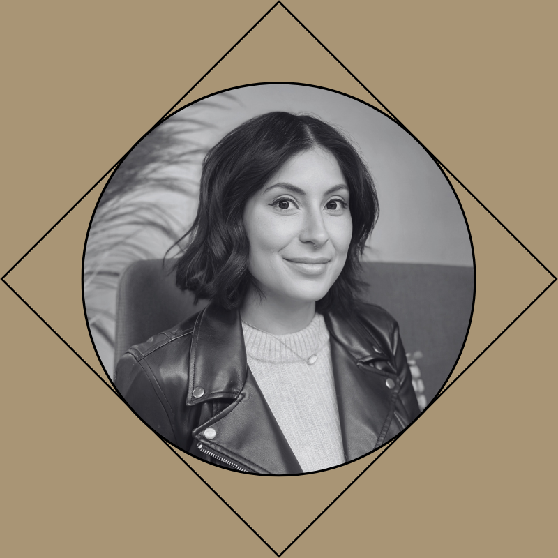 A black and white portrait of a female hairstylist with shoulder-length wavy hair, smiling, wearing a leather jacket and a light-colored sweater, sitting in a chair.