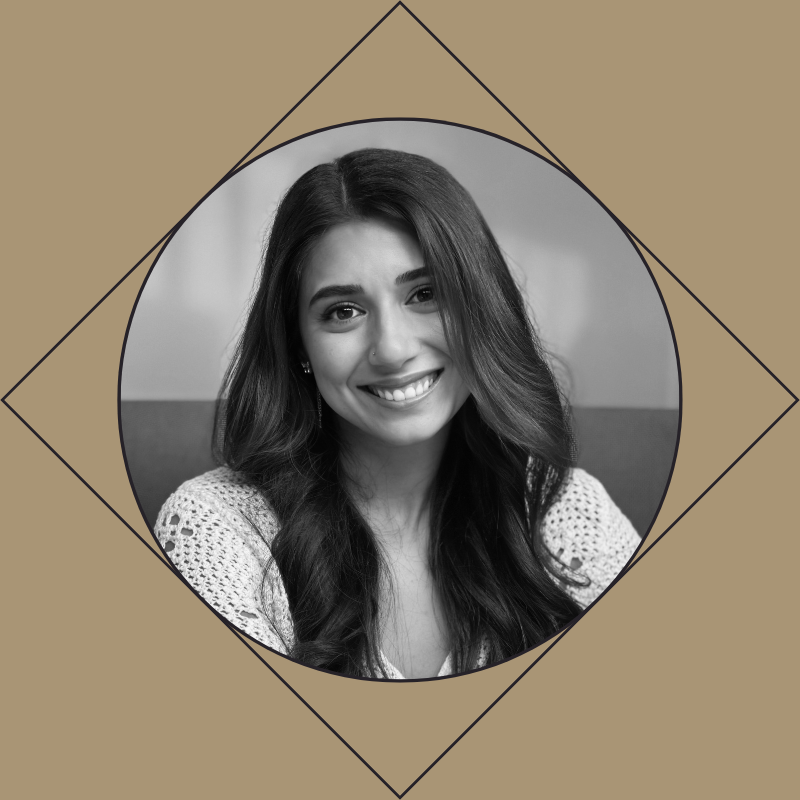 Black and white portrait of a young female hairstylist with long hair, smiling and showing her teeth, wearing a knitted top, sitting indoors.