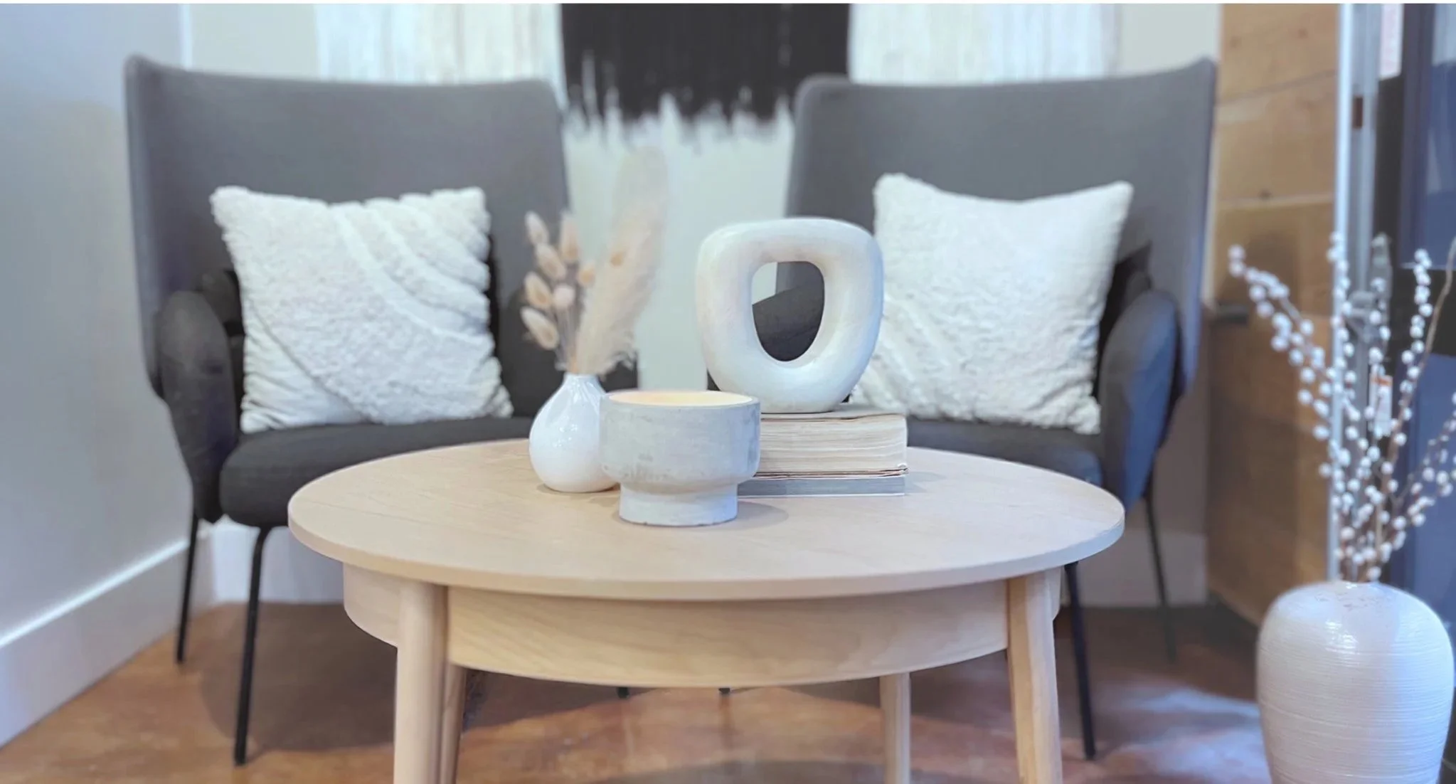 A cozy hair salon waiting area with a round wooden coffee table, two black armchairs with white textured pillows, a beige-lit vase with dried plants on the table, and a white textured vase with branches to the side.