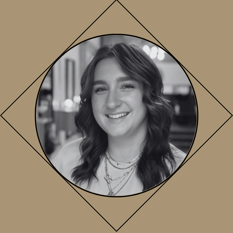 Black and white portrait of a female hairstylist with wavy hair, smiling, wearing layered necklaces, in an indoor setting.