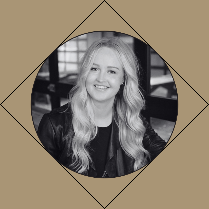 Black and white portrait of a young woman with long, wavy blonde hair, smiling, sitting indoors in front of windows.