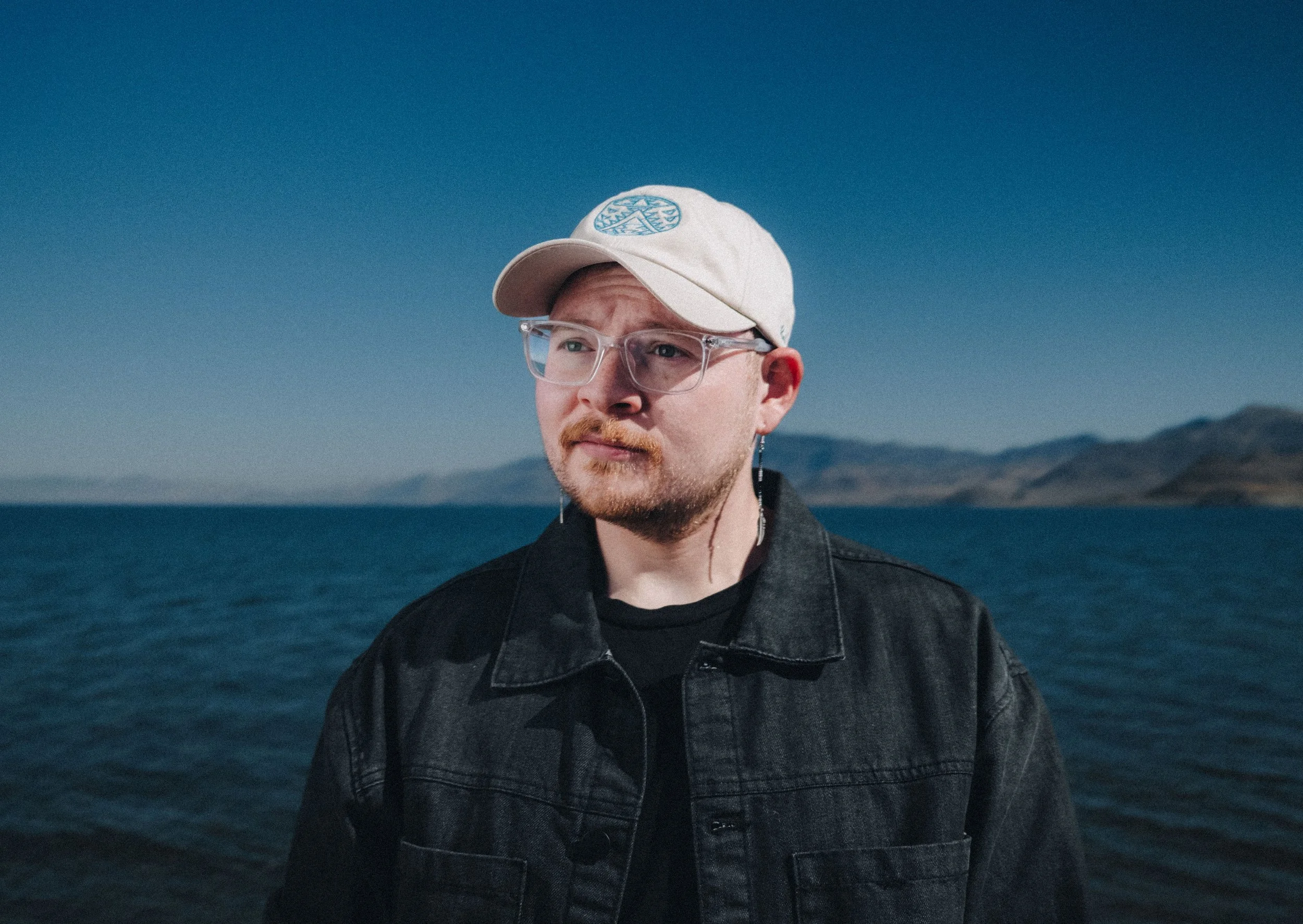 Person wearing a beige baseball cap, clear glasses, and dark jacket stands near a body of water with a mountainous background.