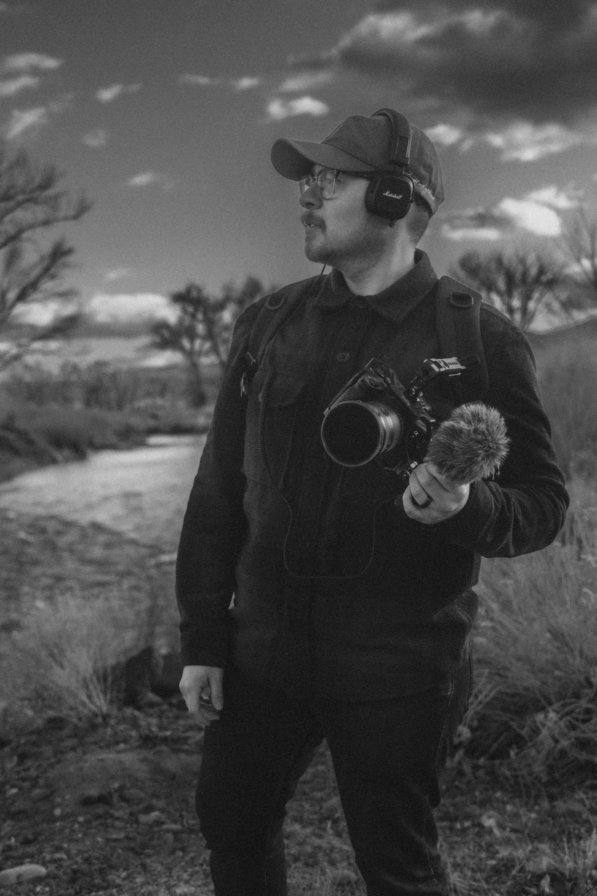 Black and white photo of a man outdoors wearing a cap and headphones, holding a camera and a microphone. He is standing near a barren landscape with trees and a cloudy sky.