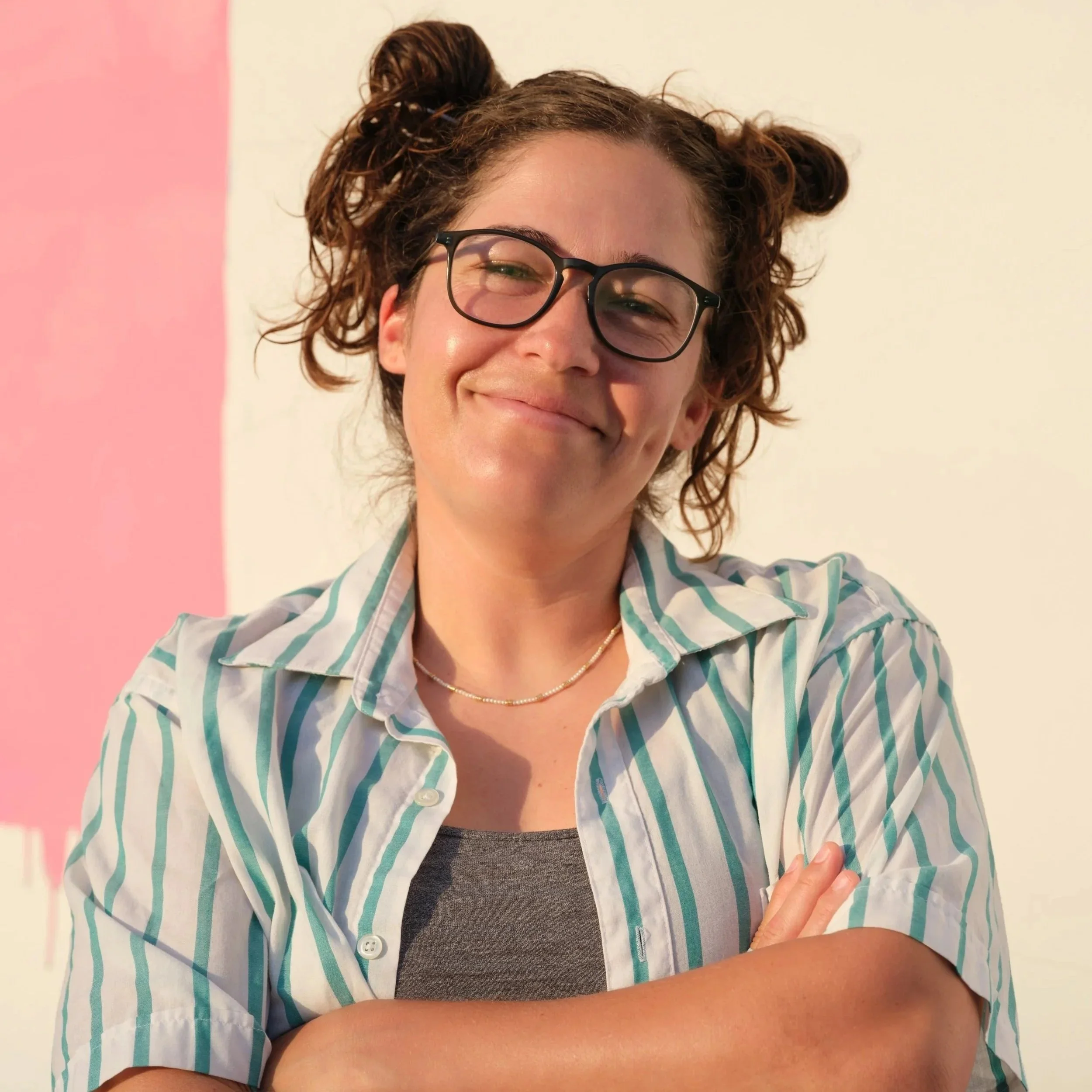 A woman with curly hair, wearing glasses, smiling, and standing against a pink and white background. She is wearing a striped shirt and has her arms crossed.
