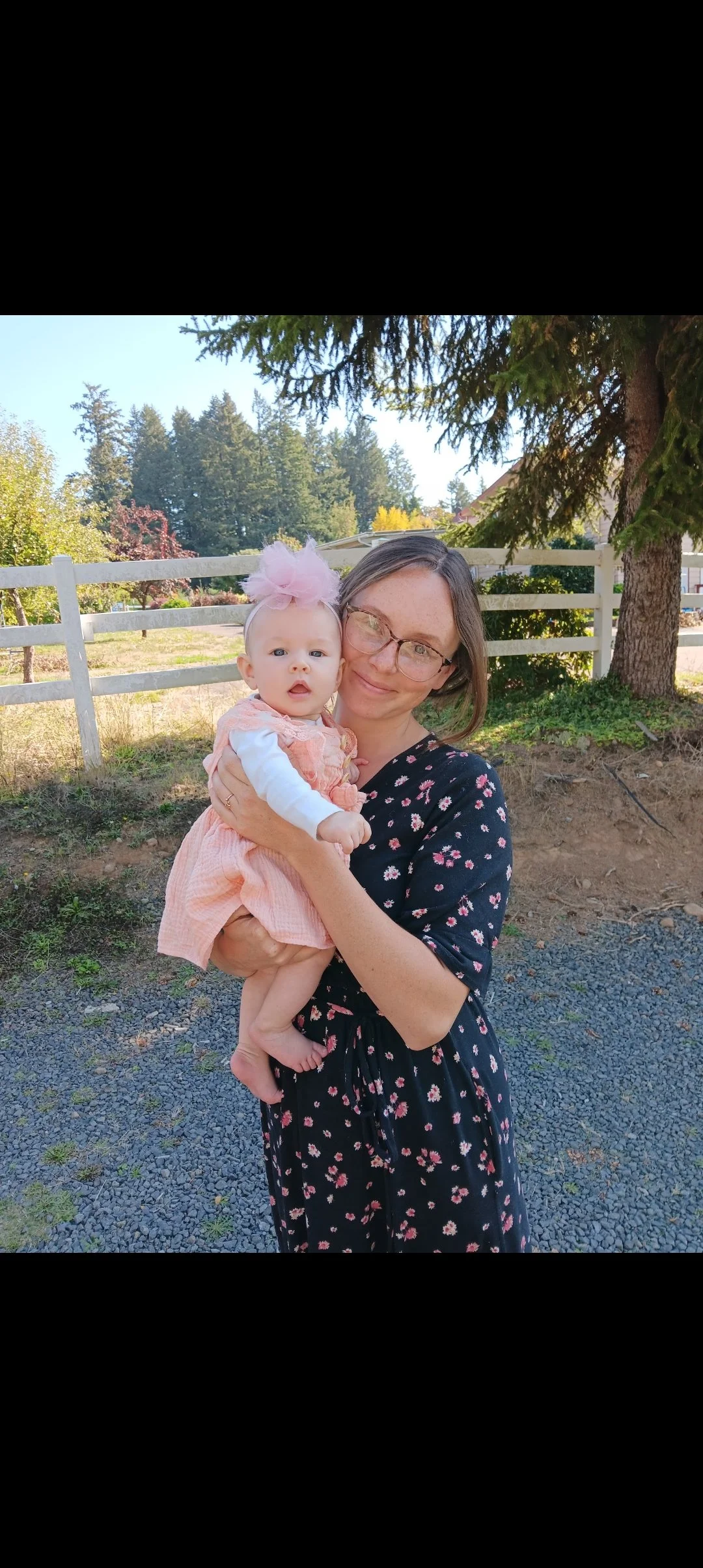 Woman wearing black floral dress and glasses smiles and holds up a baby in a pink dress and a pink bow