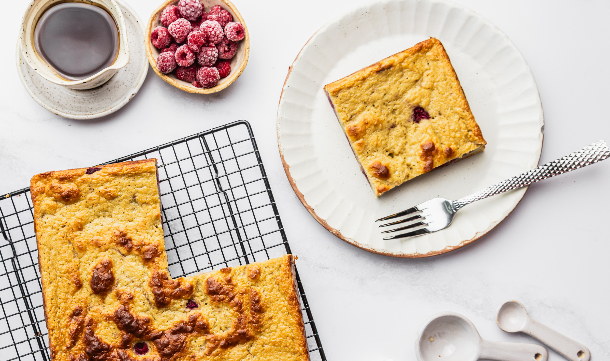 Golden baked raspberry pancake squares on a wire rack, one slice plated with fresh raspberries and coffee.