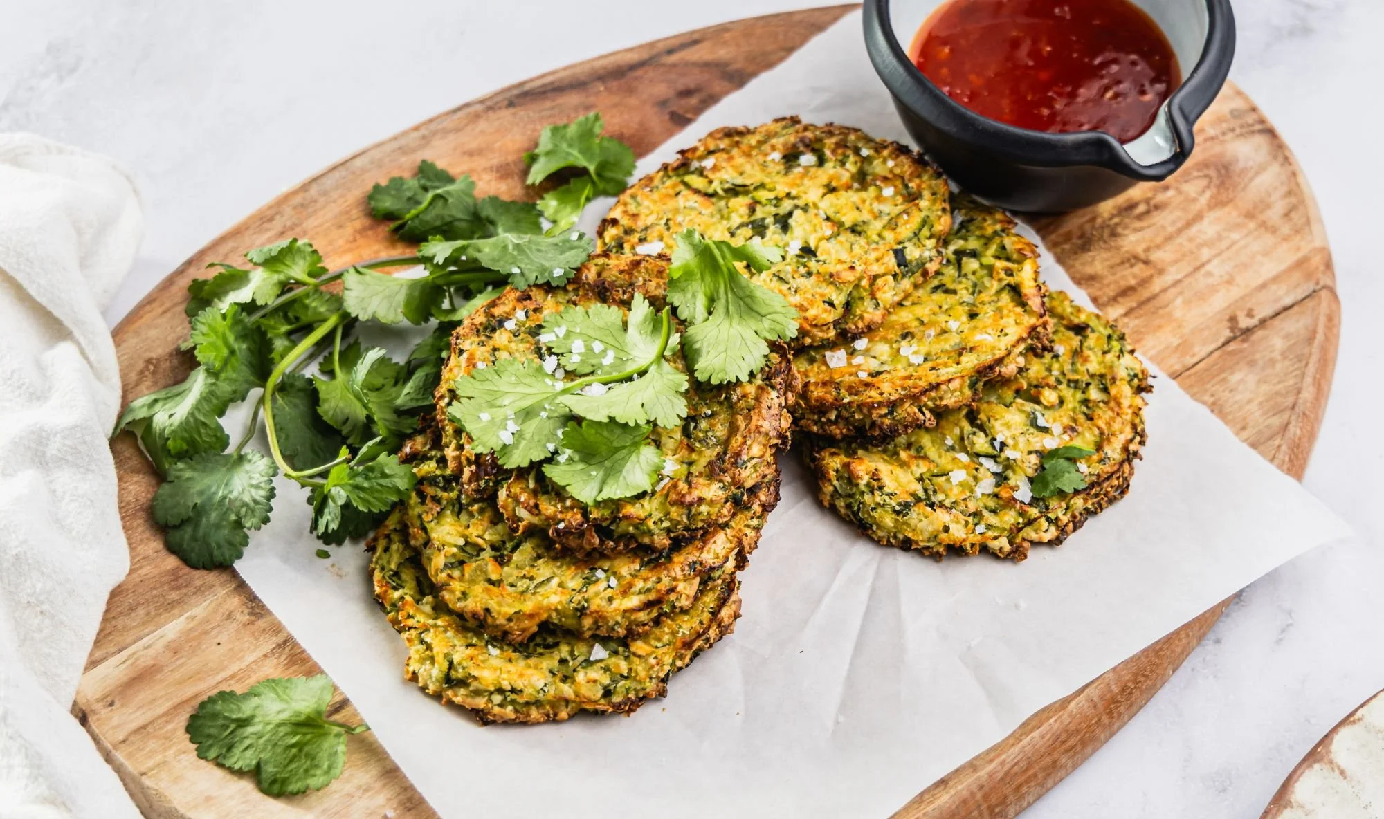 Stack of golden baked halloumi and zucchini fritters topped with fresh coriander, served on a wooden board with a bowl of sweet chilli sauce