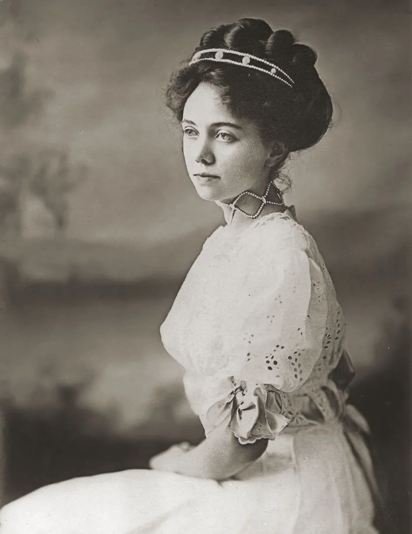 A black-and-white portrait of a young woman with styled hair, wearing a decorative headband, a choker necklace, and a dress with puffed sleeves and eyelet embroidery, looking thoughtfully to the side.