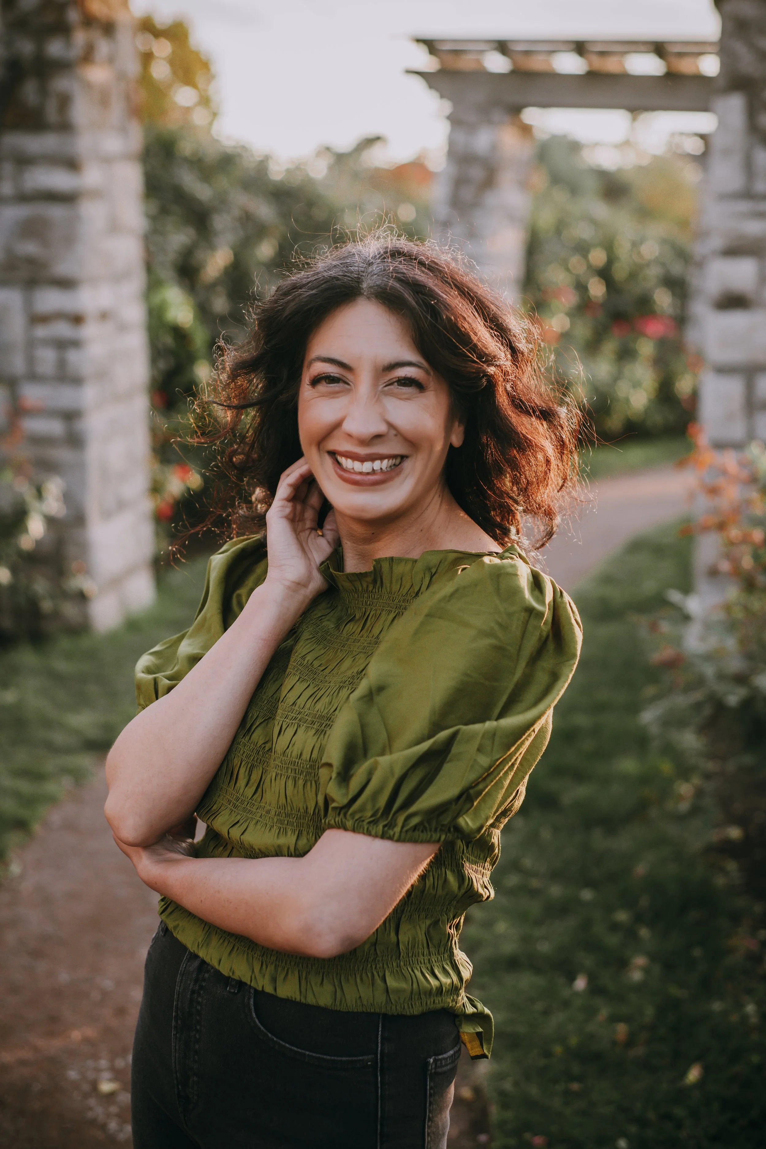 A woman with curly dark hair smiling outdoors, standing on a pathway with stone pillars and greenery in the background, wearing a green puff-sleeve blouse.