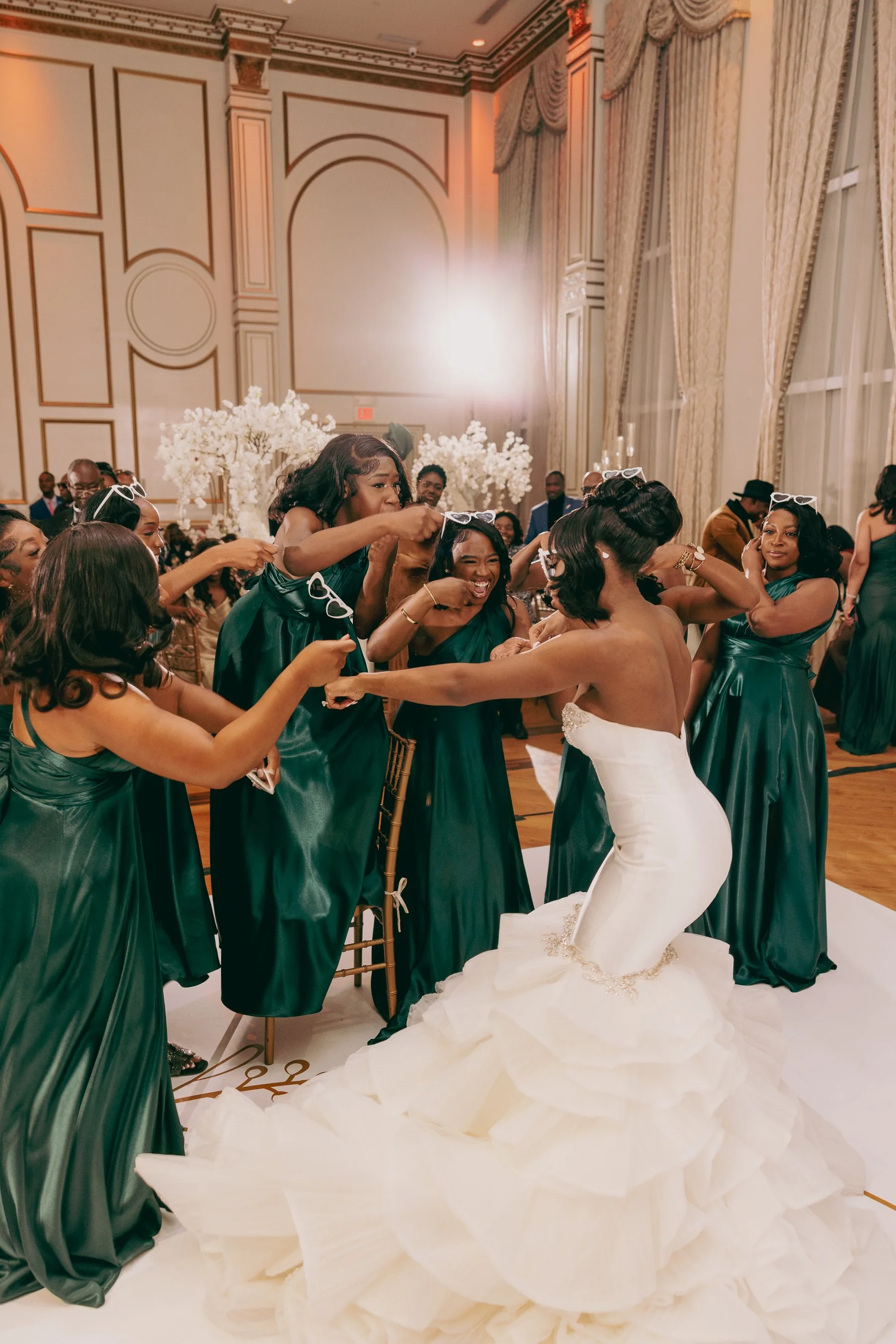 A bride in a strapless wedding gown with a ruffled train dances with her bridesmaids in teal dresses at a wedding reception in an elegant ballroom decorated with white floral arrangements.