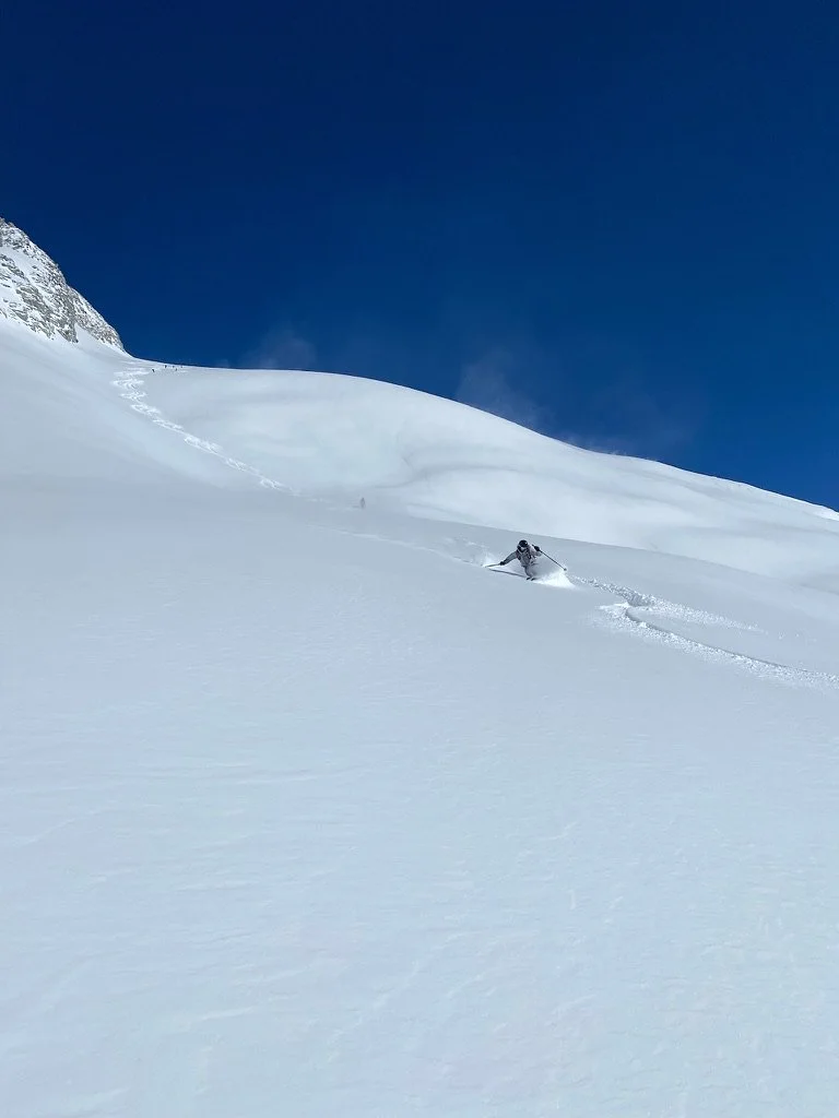 A lone skier descending an open powder face under deep blue sky — private ski guiding with Santé