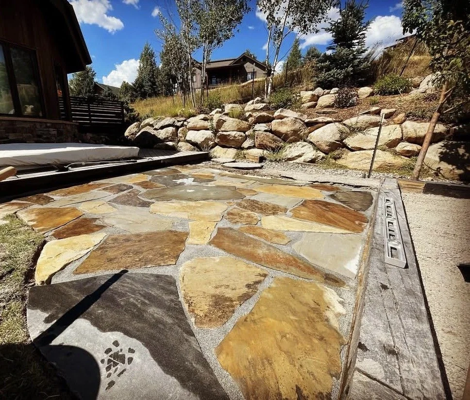 A backyard patio with a stone paved surface, a wooden house on the left, and a rock garden with trees and shrubs in the background under a partly cloudy sky.