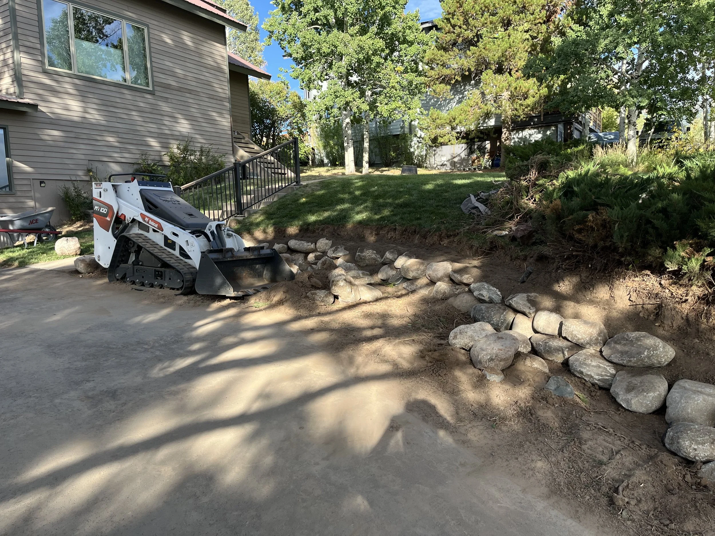 A small excavator or skid-steer loader clearing a patch of dirt next to a house with a lawn and trees.