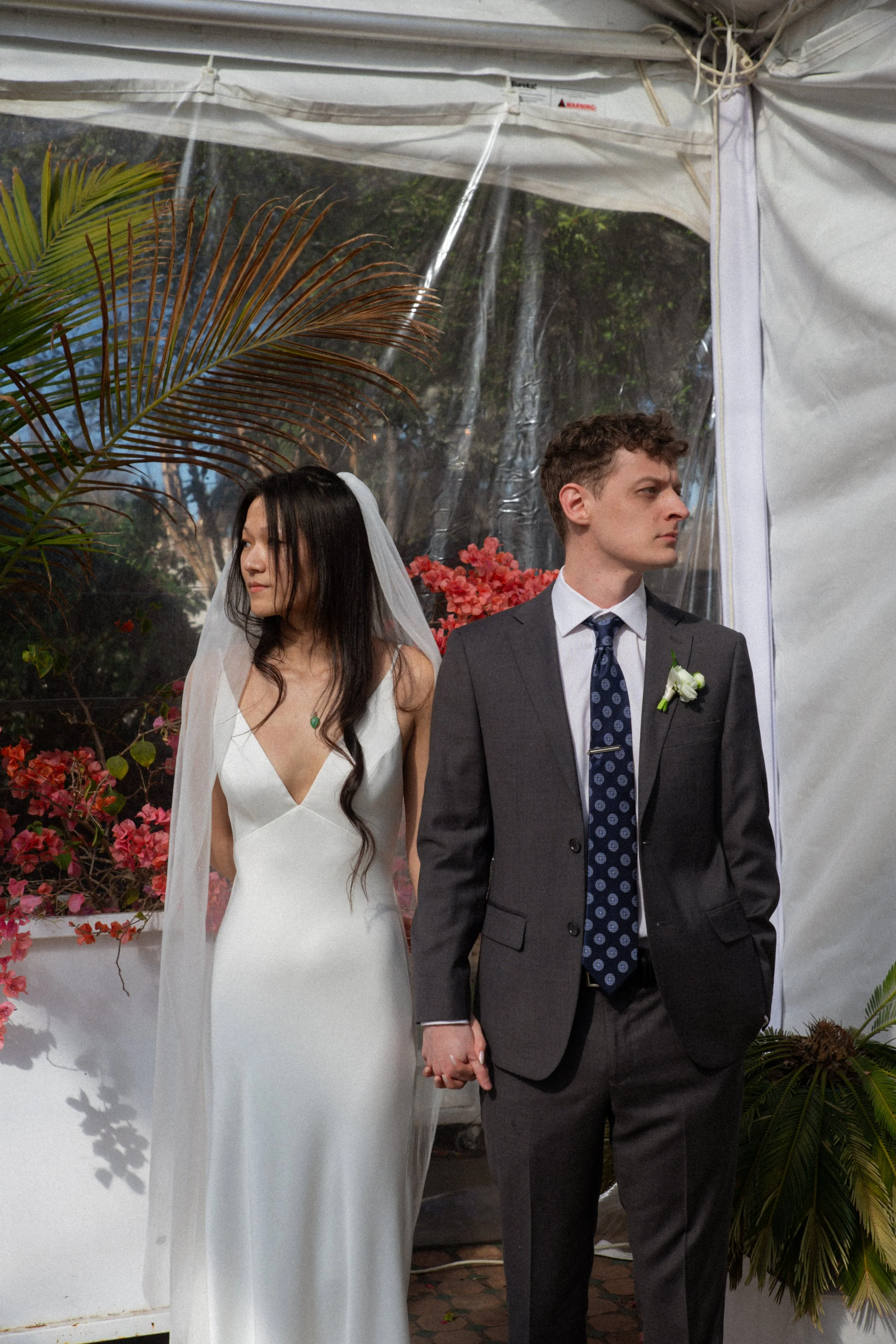 A bride and groom holding hands at their wedding, standing inside a tent with floral and green foliage decorations.