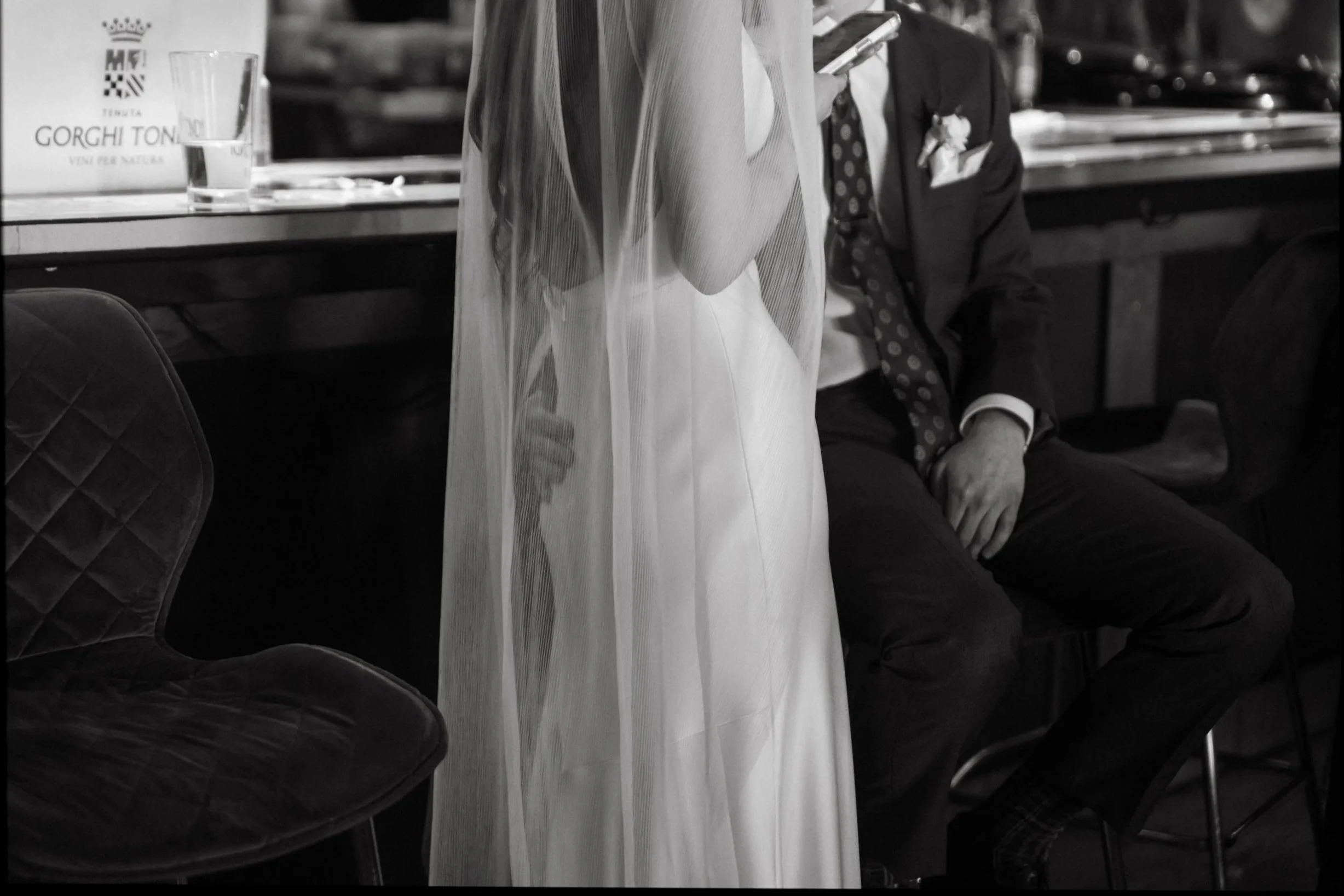 A person in a suit and tie sitting on a chair, looking at their phone, partially obscured by a transparent curtain, with a table and a box labeled 'Gorgi Ton' in the background.