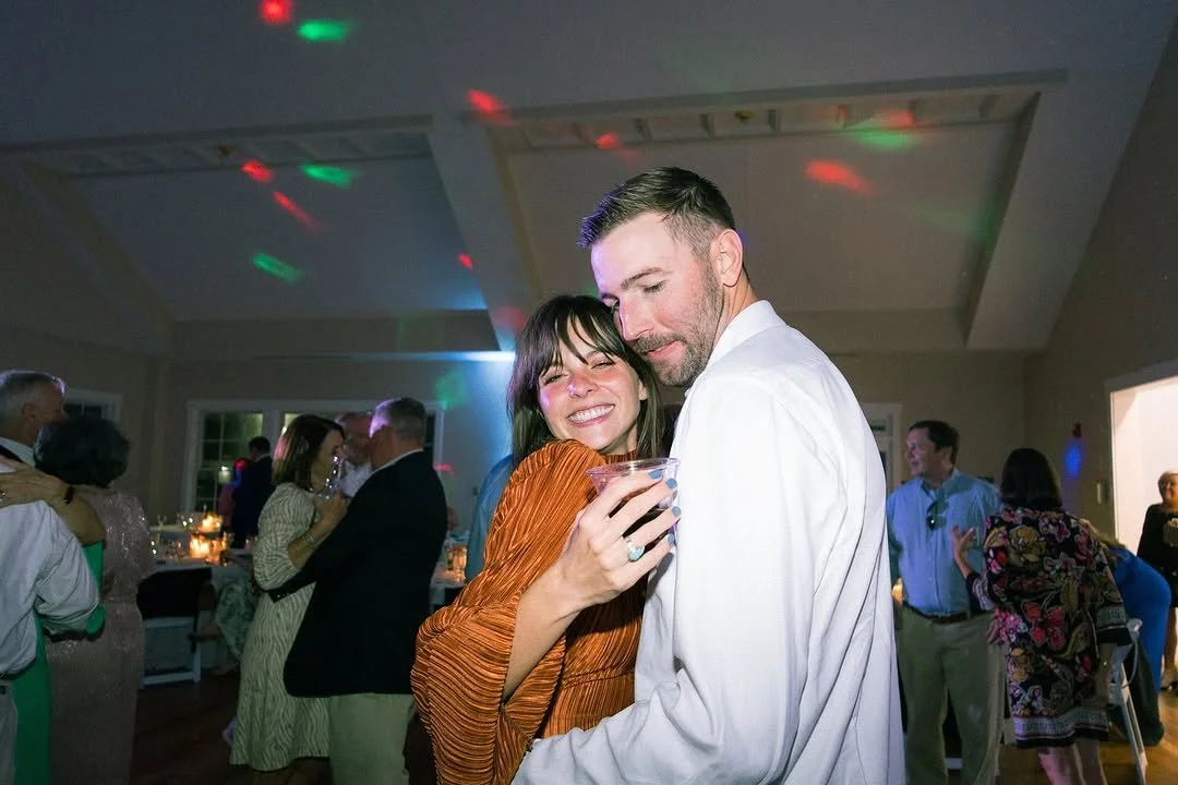 A woman with dark hair and a man with a short beard at a party or celebration, holding a drink, with other people dancing and socializing in the background.
