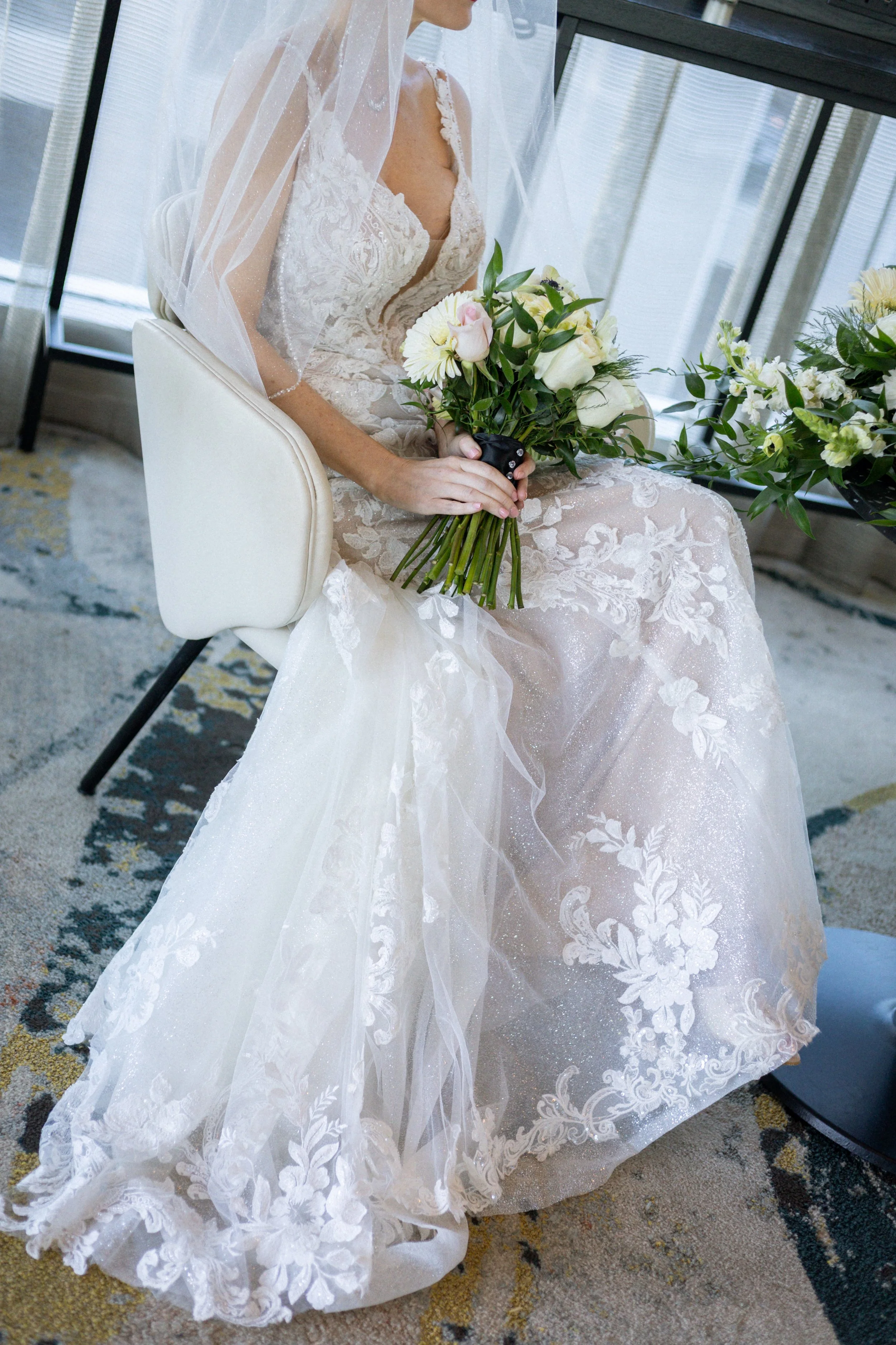 A bride sitting in a white chair holding a bouquet of pink and white roses, wearing a detailed lace wedding gown with an attached veil.