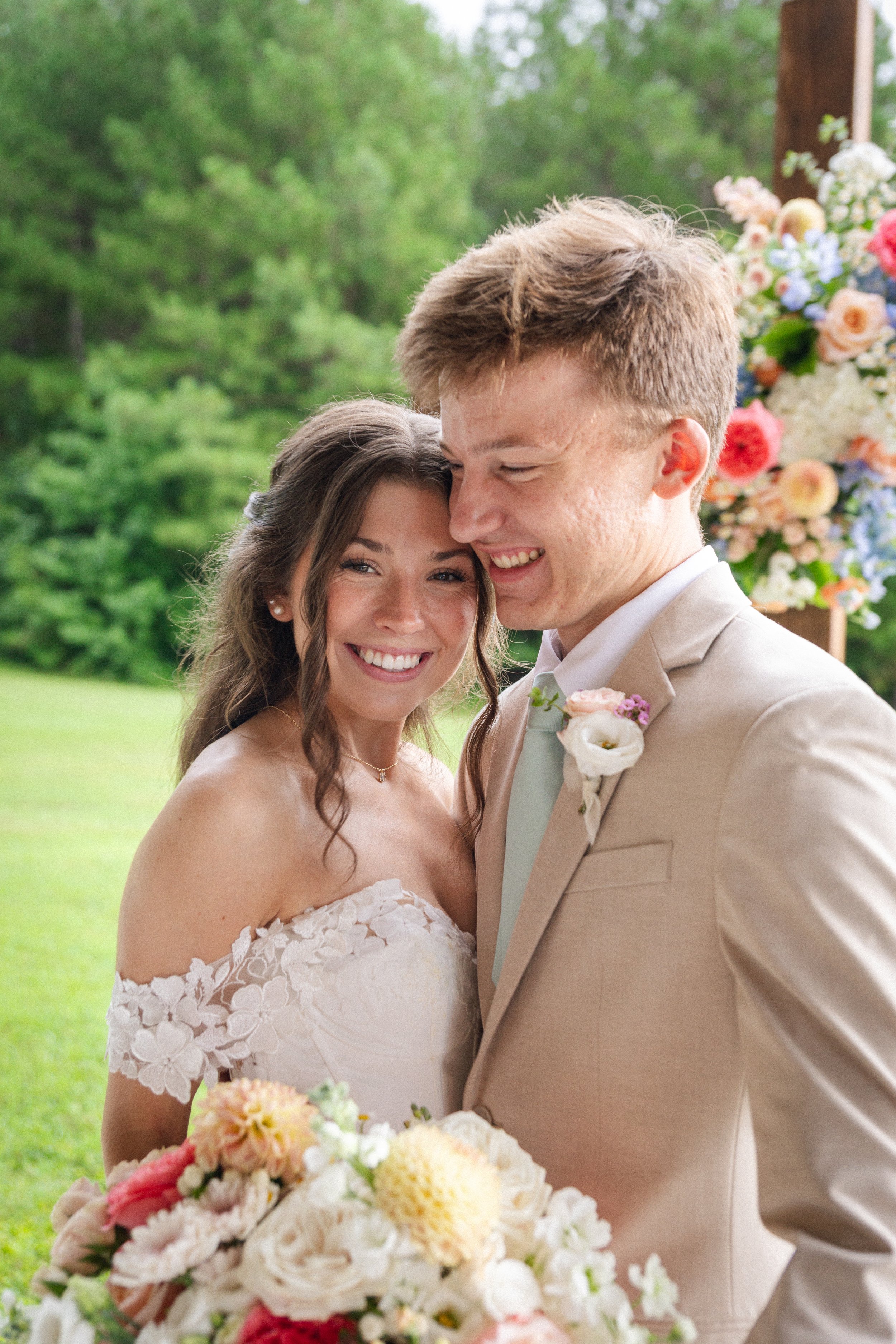 A smiling bride and groom embrace outdoors during a wedding, with green trees and a floral arrangement in the background.