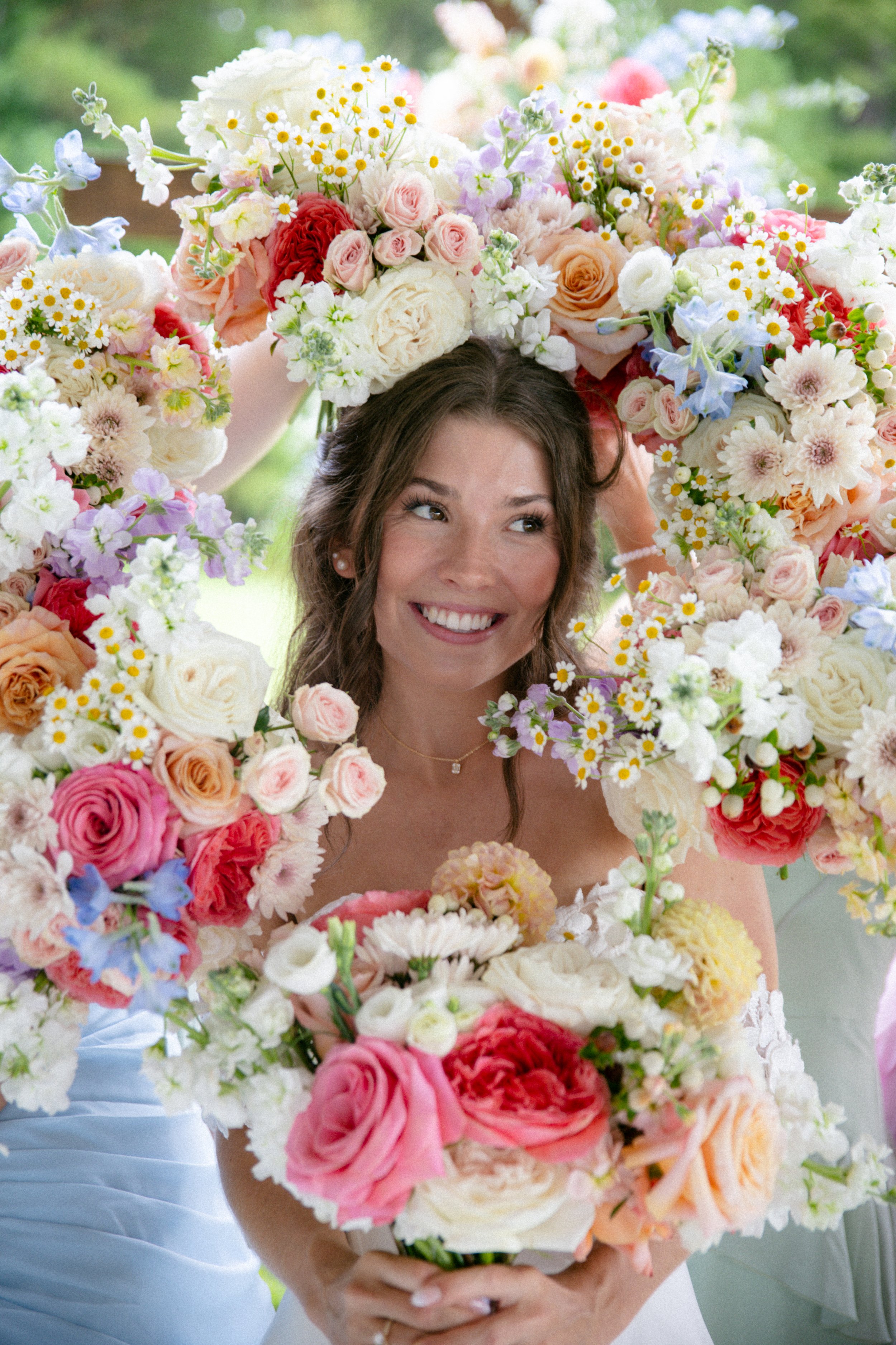 Bride smiling through a colorful floral wreath surrounded by an arrangement of assorted flowers in pastel and bright colors.