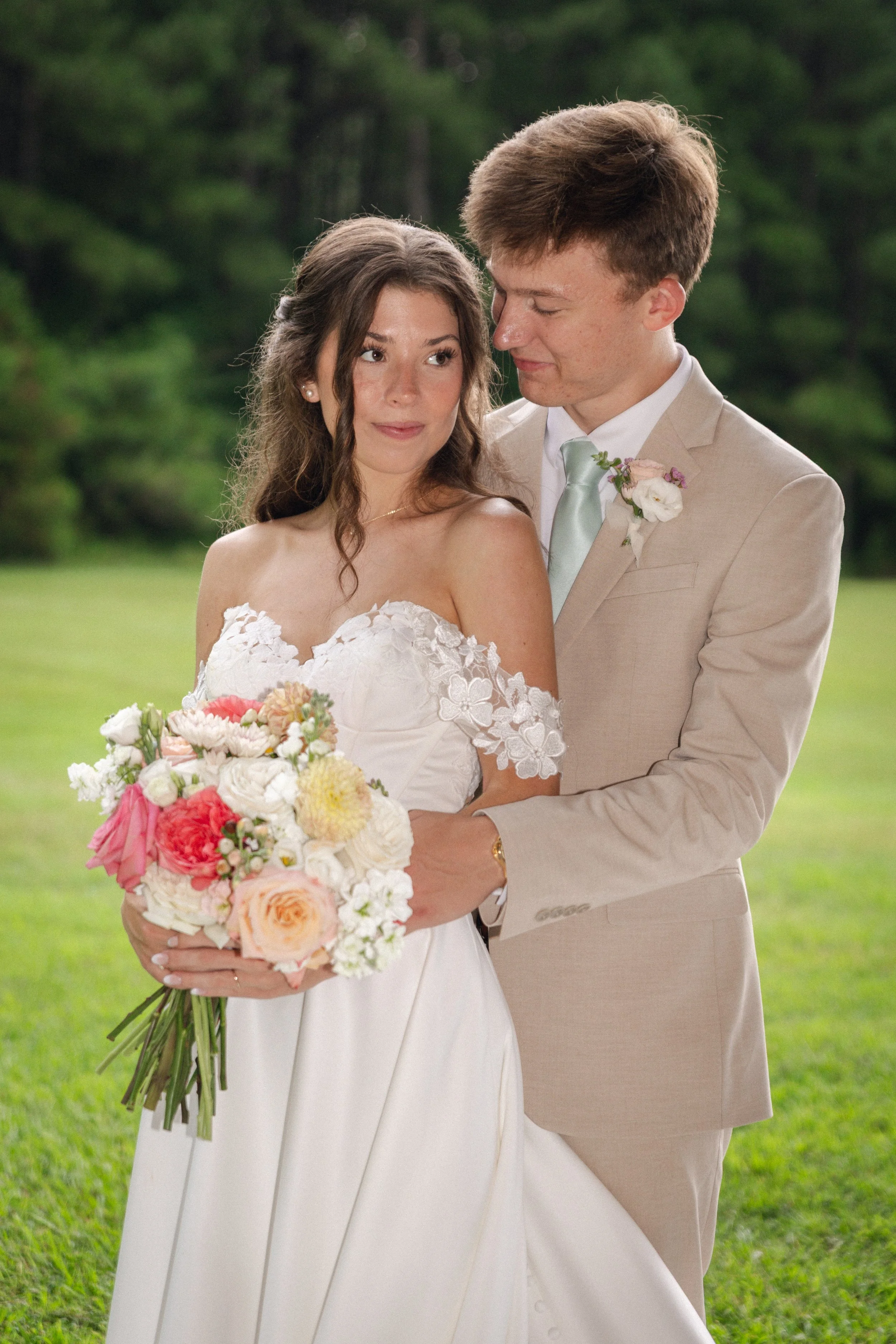 A bride in a white wedding dress holding a bouquet of pink, yellow, and white flowers, standing next to her groom in a beige suit with a light blue tie, outdoors with green trees and grass in the background.