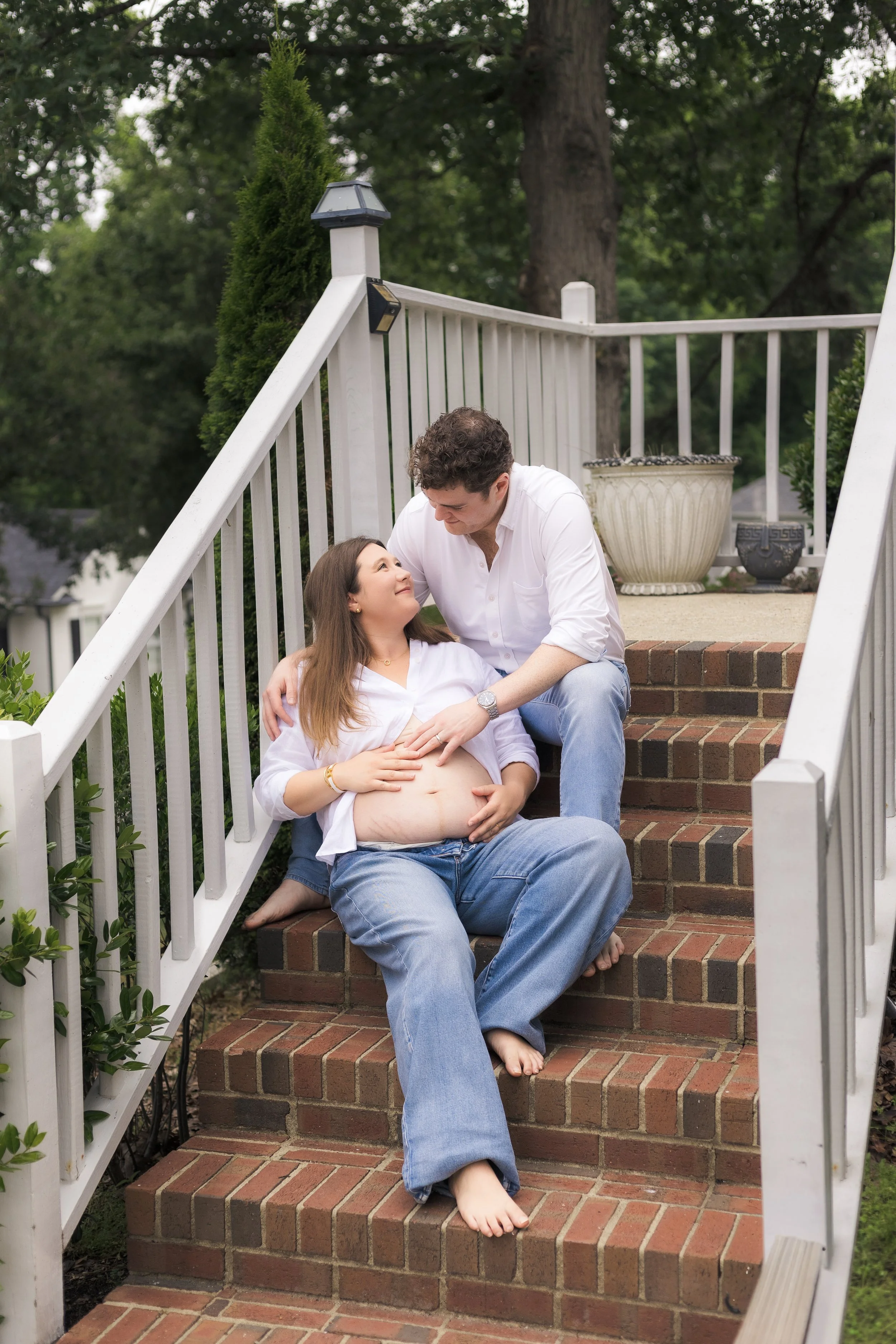 A man and a woman sitting on brick stairs outside. The woman is pregnant and lying back, with the man beside her. They are looking at each other affectionately. The setting is in a green, leafy area with a white fence and large decorative pots nearby.