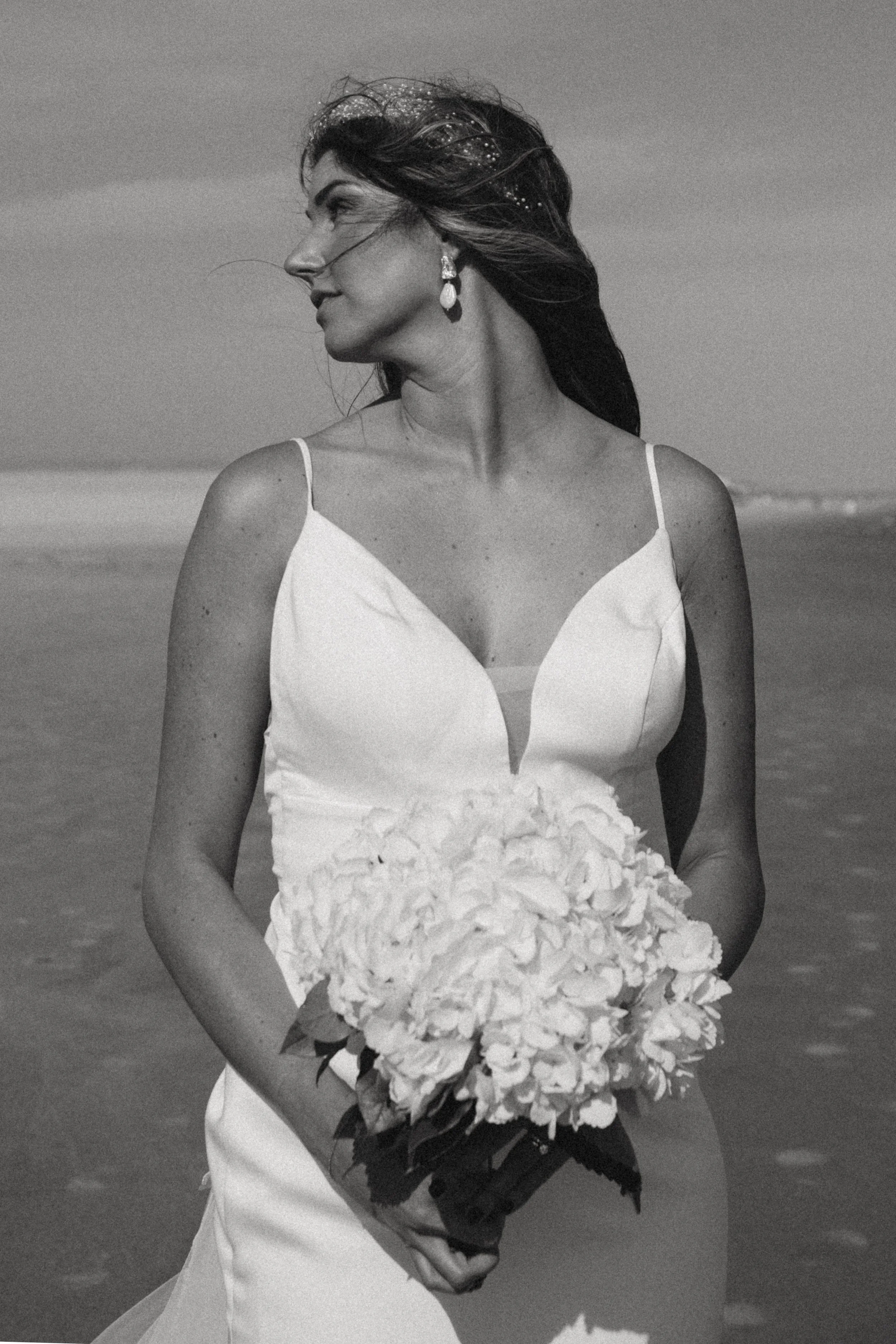 A woman in a white dress holding a bouquet of white flowers at the beach, with wind blowing her hair, photographed in black and white.