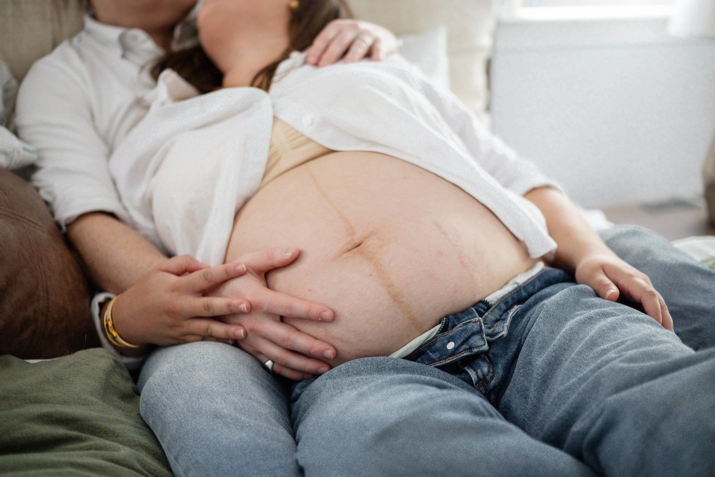 A couple sitting on a bed, with the woman resting her head on the man's chest. The woman is pregnant, and her bare belly is visible.