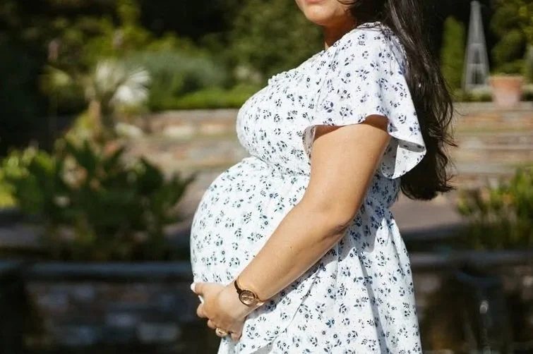 A pregnant woman wearing a white floral dress standing outdoors, cradling her belly.