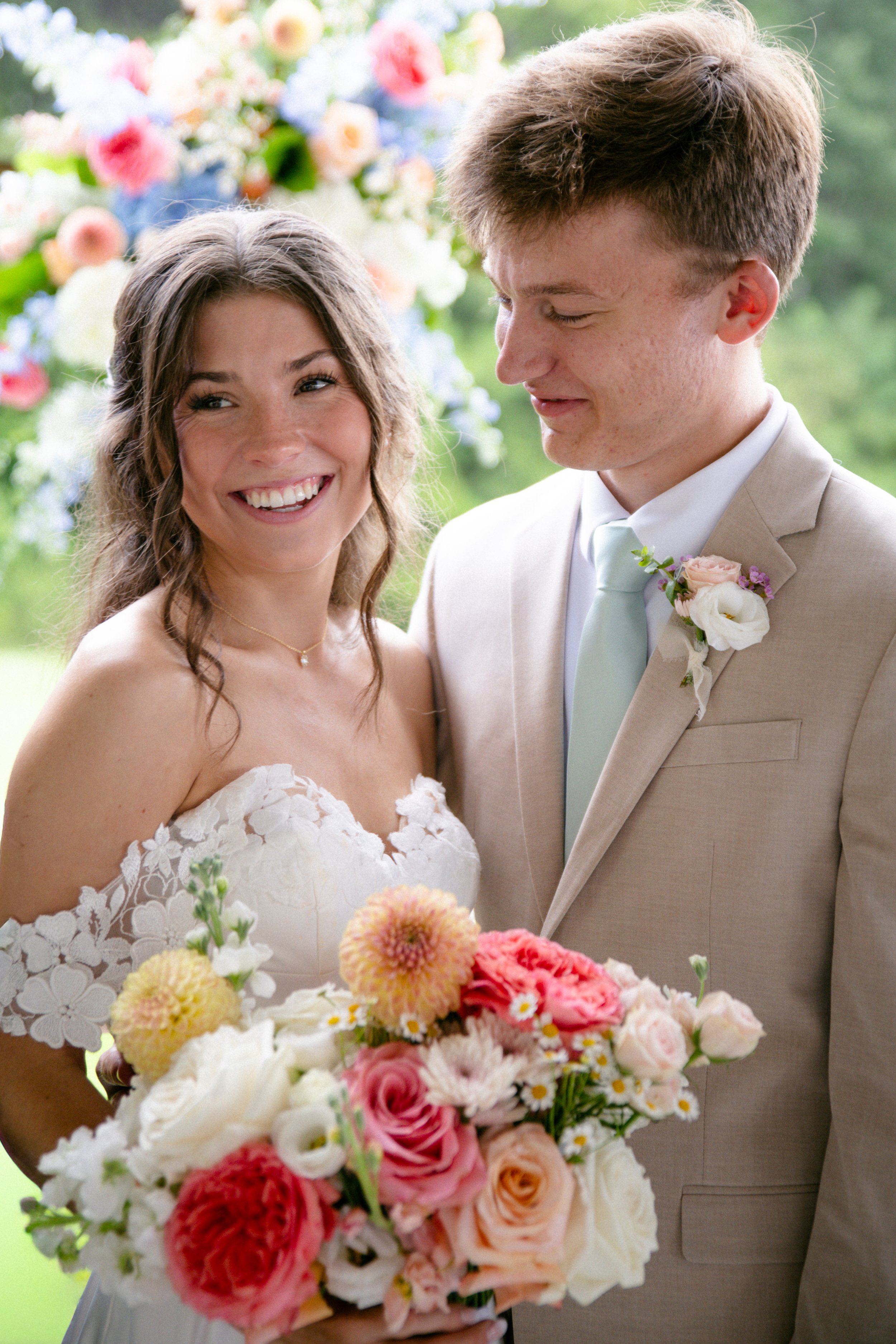 A bride and groom smiling at each other during their wedding, with a colorful bouquet of flowers in the bride's hands and a floral backdrop in the background.