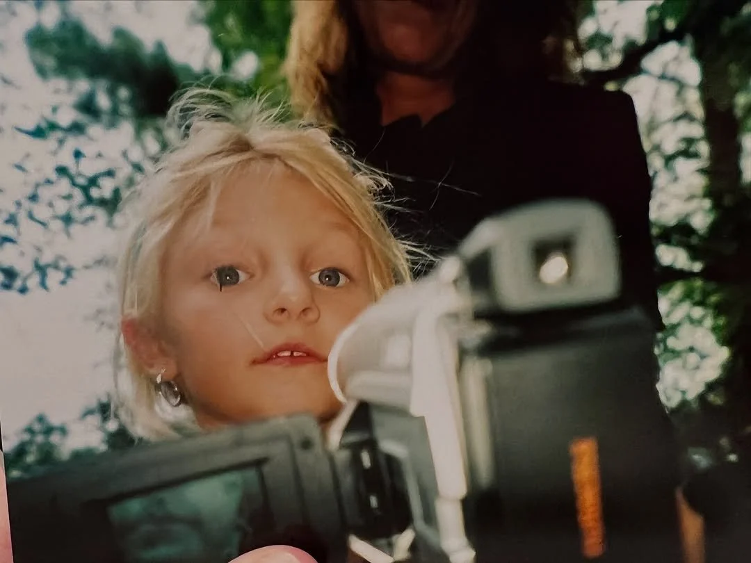 A young girl with blonde hair looking into the camera while being filmed or photographed, with a woman standing behind her outdoors.
