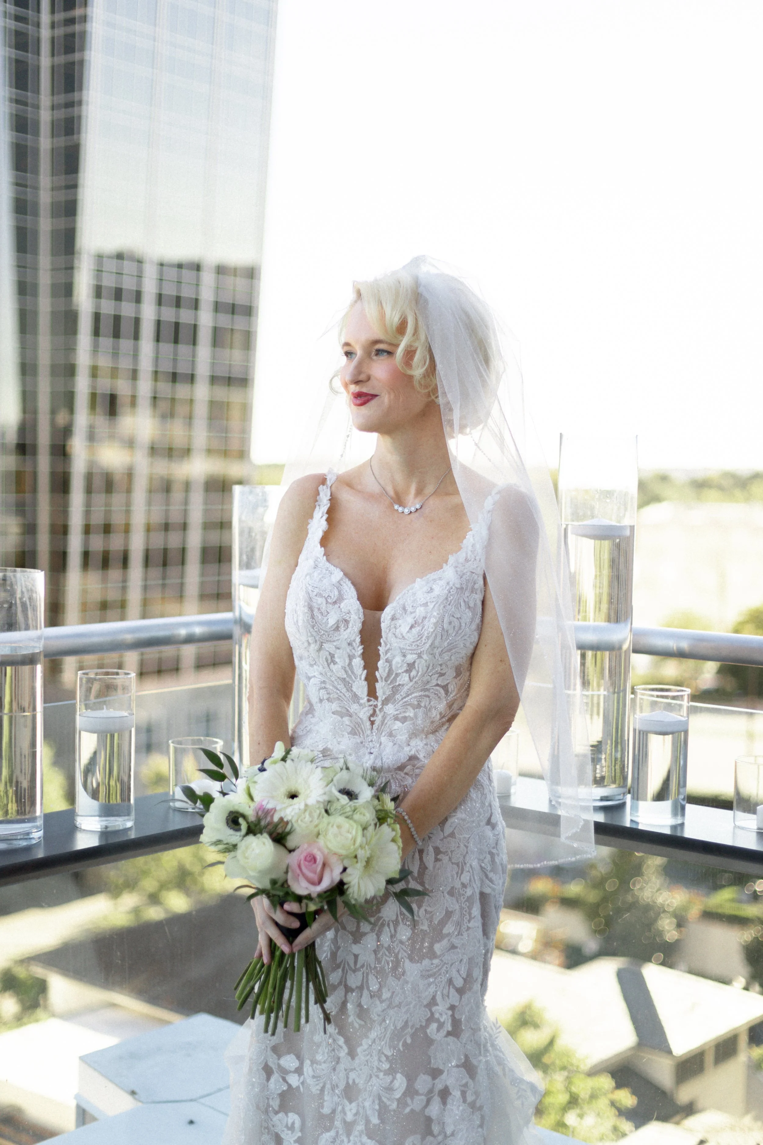 A bride with blonde hair wearing a white lace wedding dress and a veil, holding a bouquet of white and pink flowers, standing outdoors on a balcony with glass of water and modern building in background.