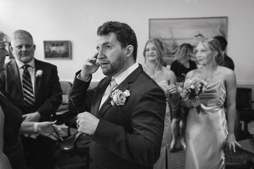 Black and white photo of a wedding reception with a groom talking on the phone, surrounded by guests including a woman holding a bouquet of flowers.